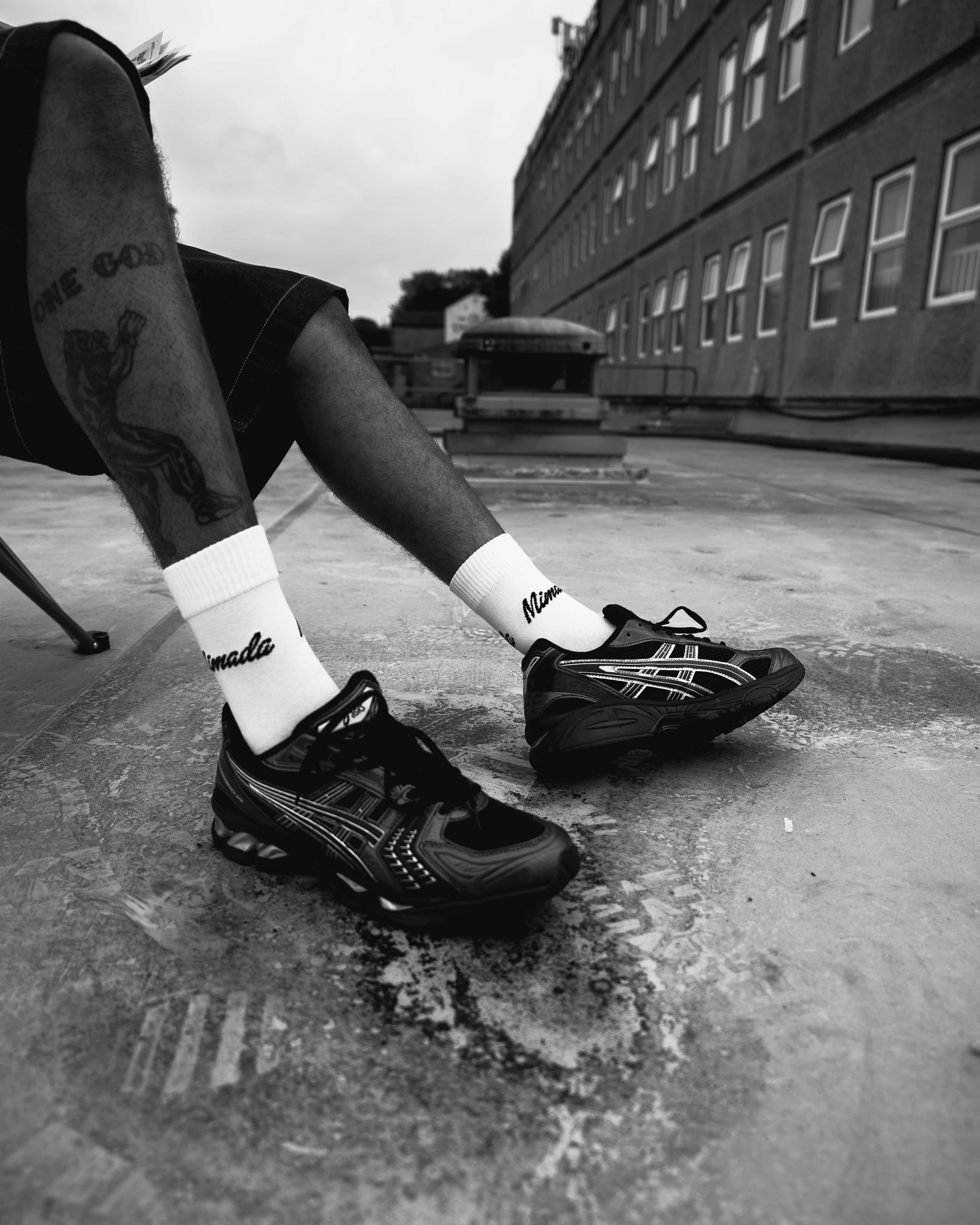 Person wearing black sneakers and white socks with a brand logo, standing on a rooftop.
